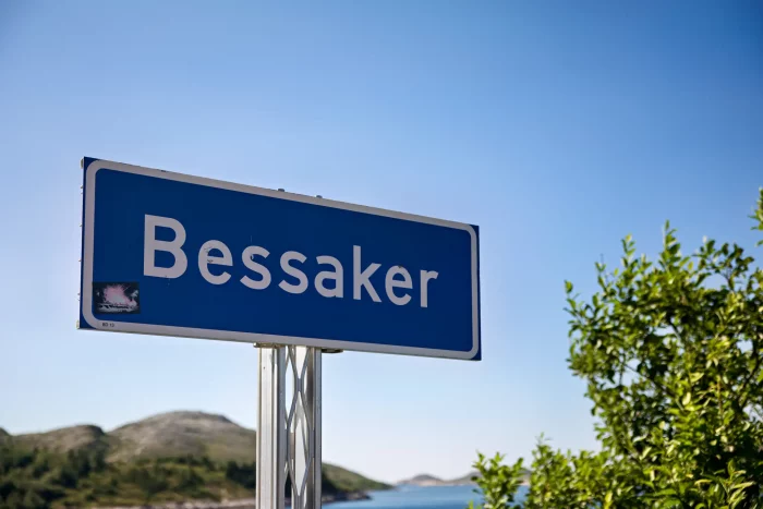 Straßenschild mit dem Ortsnamen 'Bessaker' vor klarem Himmel und Landschaft mit Hügeln und Wasser