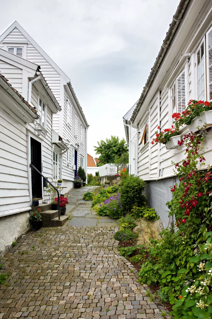 Schmaler gepflasterter Weg zwischen weißen Holzhäusern mit Blumen in Blumenkästen und Gärten