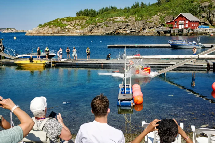 Menschen beobachten ein Kajakrennen auf einem See mit einer roten Hütte und bewaldeten Felsen im Hintergrund