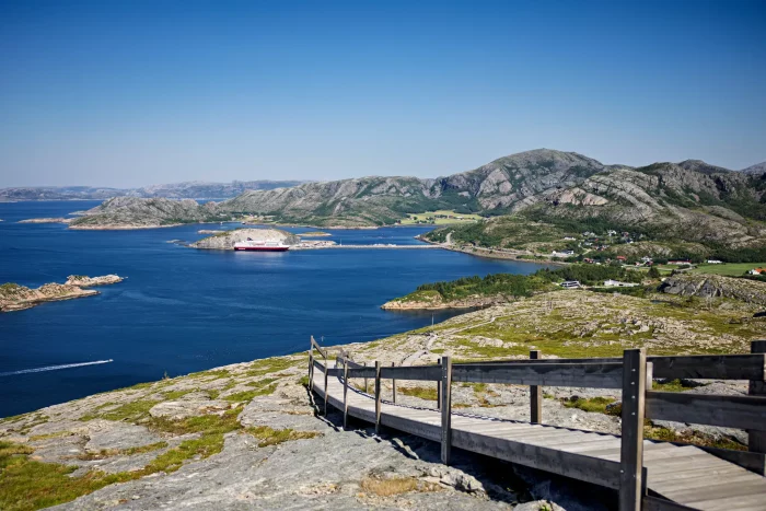 Blick auf einen Fjord mit einem Hurtigruten-Kreuzfahrtschiff, umgeben von felsigen Inseln und grüner Landschaft unter klarem Himmel
