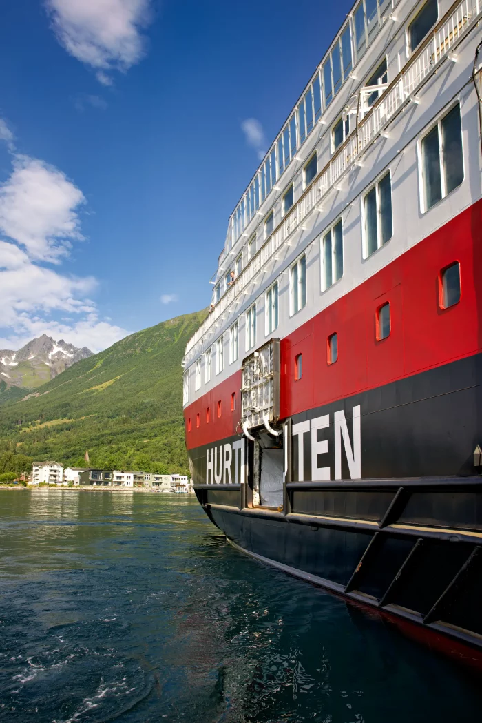 Hurtigruten-Kreuzfahrtschiff am Wasser mit Bergen und Häusern im Hintergrund unter blauem Himmel