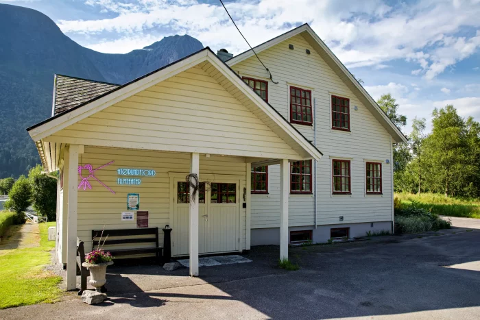 Weißes, zweistöckiges Haus mit rotem Fensterrahmen und überdachtem Eingang in ländlicher Umgebung mit Bergen im Hintergrund
