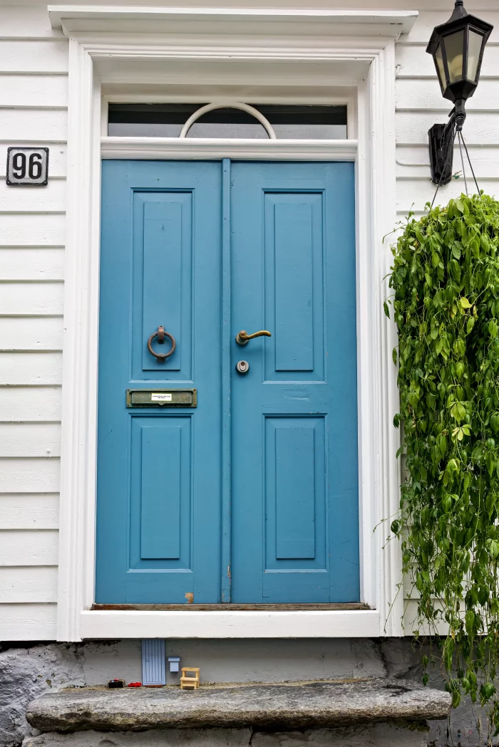 Blaue Doppeltür mit Briefkasten und Türklopfer an weißem Haus, daneben Laterne und hängende grüne Pflanze