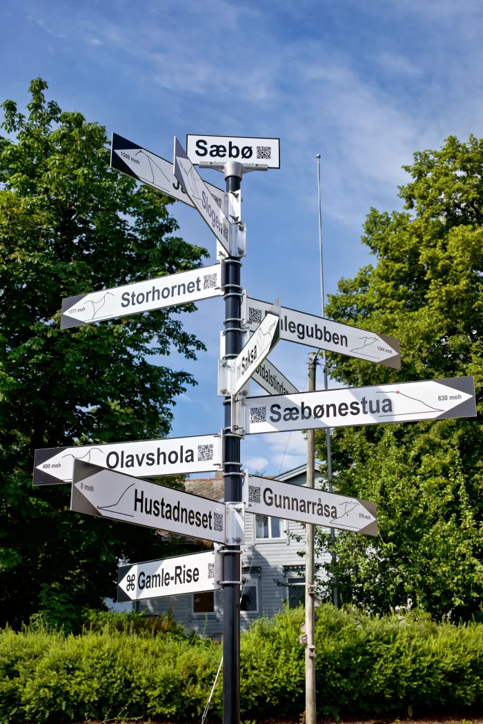 Wegweiser mit mehreren Schildern zu Orten wie Sæbø, Storhornet, Olavshola und Hustadneset vor grünem Laub und blauem Himmel