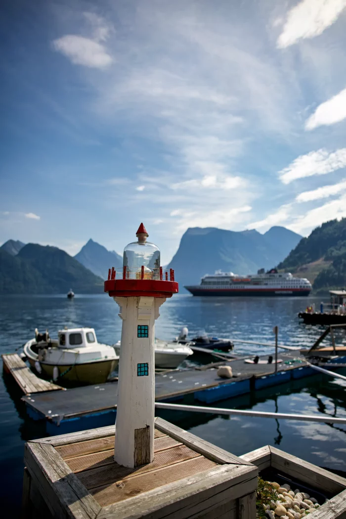 Modell eines weißen Leuchtturms mit roter Spitze am Hafen, dahinter Hurtigruten-Kreuzfahrtschiff auf Fjord mit Bergen im Hintergrund