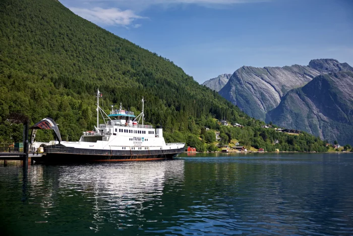 Fähre am Kai vor bewaldeten Bergen und steilen Felsformationen unter blauem Himmel