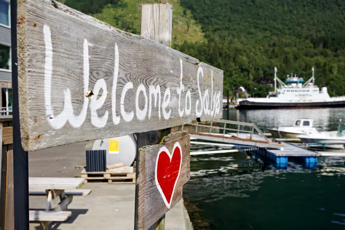 Holzschild mit weißer Schrift 'Welcome to Sabo' und rotem Herz, Hafen mit Booten und grünem Wald im Hintergrund