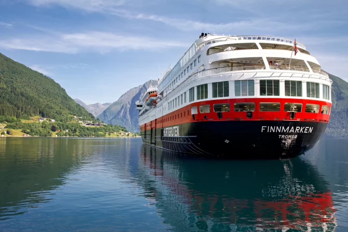 Das Kreuzfahrtschiff Finnmarken der Hurtigruten-Reederei fährt in einem norwegischen Fjord, umgeben von grünen Bergen und klarem Himmel.