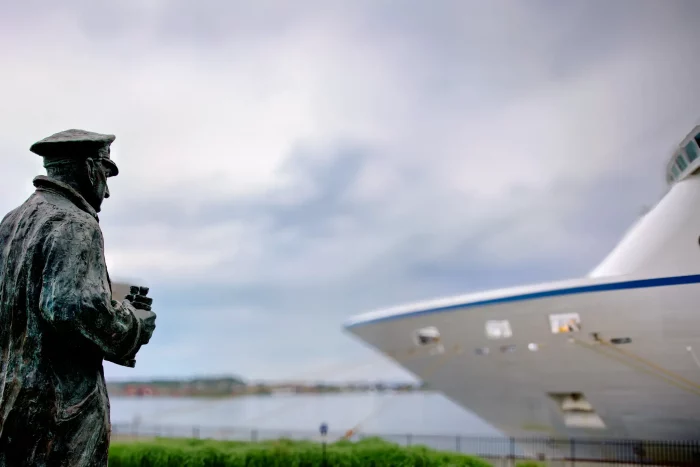 Bronzestatue eines Mannes mit Mütze und Fernglas blickt auf den Bug eines großen Kreuzfahrtschiffs am Hafen