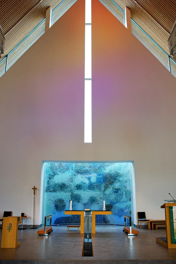 Innenraum einer modernen Kirche mit hohem, spitz zulaufendem Dach, zentralem Altar und großem Fenster mit abstraktem Muster