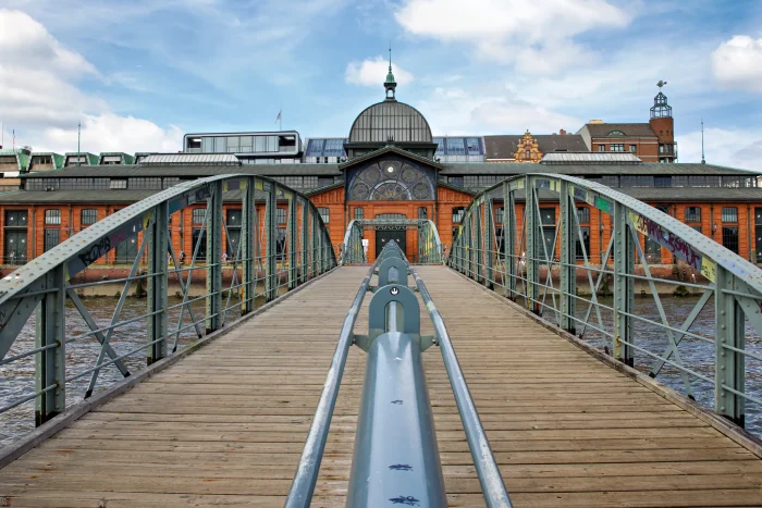 Holzsteg mit Metallgeländer führt zu einem historischen Backsteingebäude mit Kuppel und Uhr