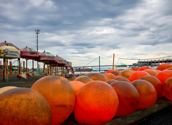 Große orangefarbene Kugeln am Hafen mit einem Karussell und einer Brücke im Hintergrund unter bewölktem Himmel