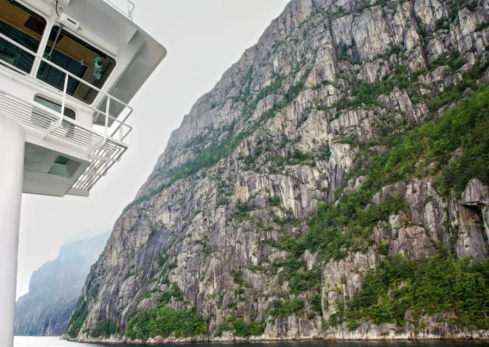 Blick von einem Schiff auf steile Felswände mit vereinzeltem Grün entlang eines Fjords
