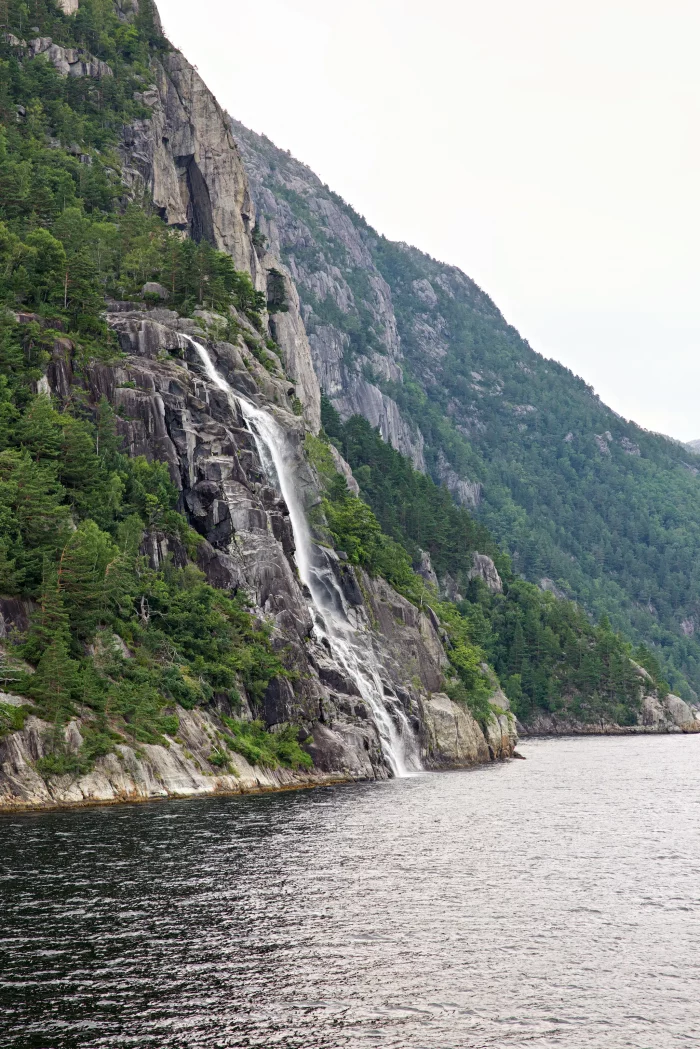 Wasserfall fließt an steiler Felswand mit grünen Bäumen in einen Fjord