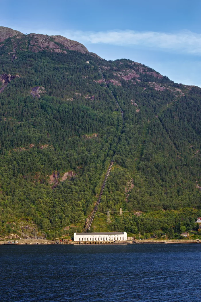 Berg mit dichter Bewaldung und einer Seilbahnlinie, die zu einem weißen Gebäude am Wasser führt