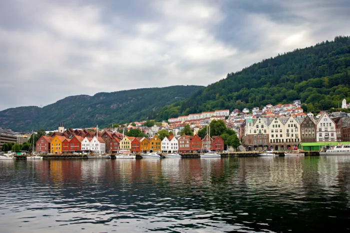 Bunte Holzhäuser am Wasser in Bergen mit bewaldeten Hügeln im Hintergrund unter bewölktem Himmel