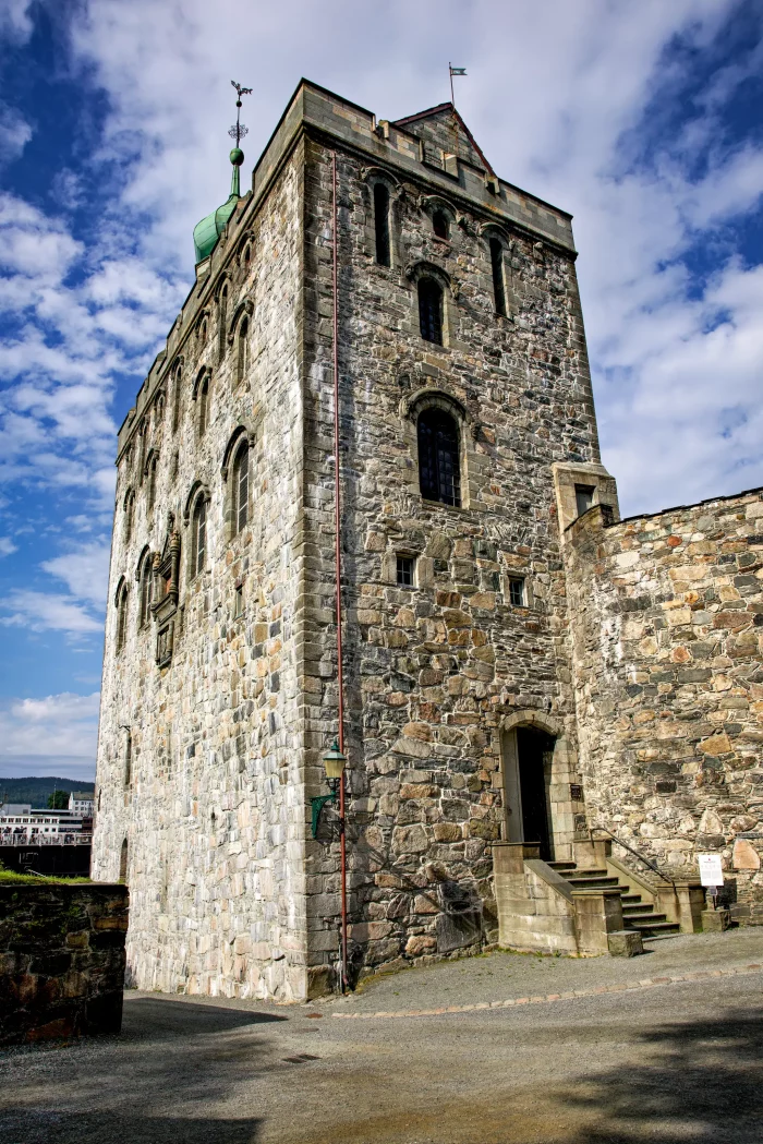 Hoher mittelalterlicher Steinturm mit kleinen Fenstern und Wetterfahne unter bewölktem Himmel