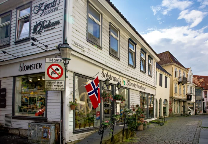 Straßenecke mit norwegischer Flagge und Geschäften, darunter 'Solstrunds Blomsterforretning' und 'Kjerstis Kjøkken' in einem historischen Holzhaus