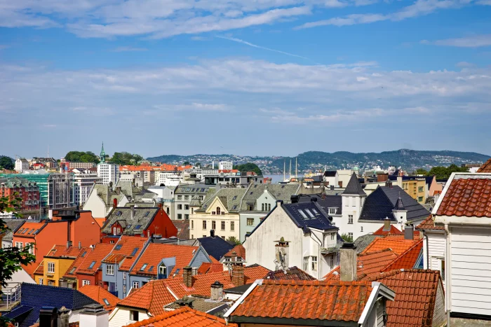 Blick über die Dächer einer Stadt mit roten Ziegeln, weißen und bunten Häusern, im Hintergrund Hügel und blauer Himmel mit Wolken