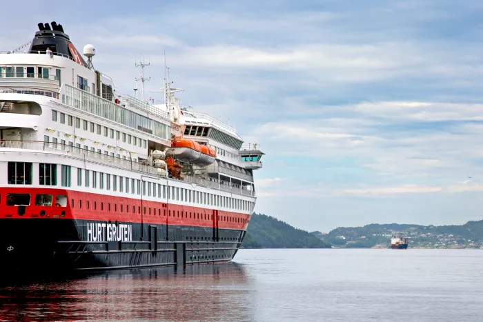 Hurtigruten-Kreuzfahrtschiff mit weißem und rotem Rumpf auf ruhigem Wasser, Küstenlandschaft im Hintergrund