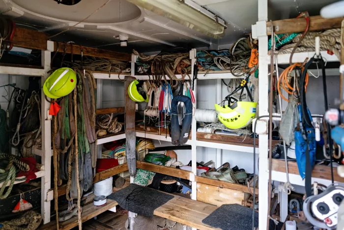 Innenraum eines Segelschiffs mit Regalen voller Seile, zwei gelben Helmen, Sicherheitsausrüstung und Werkzeug