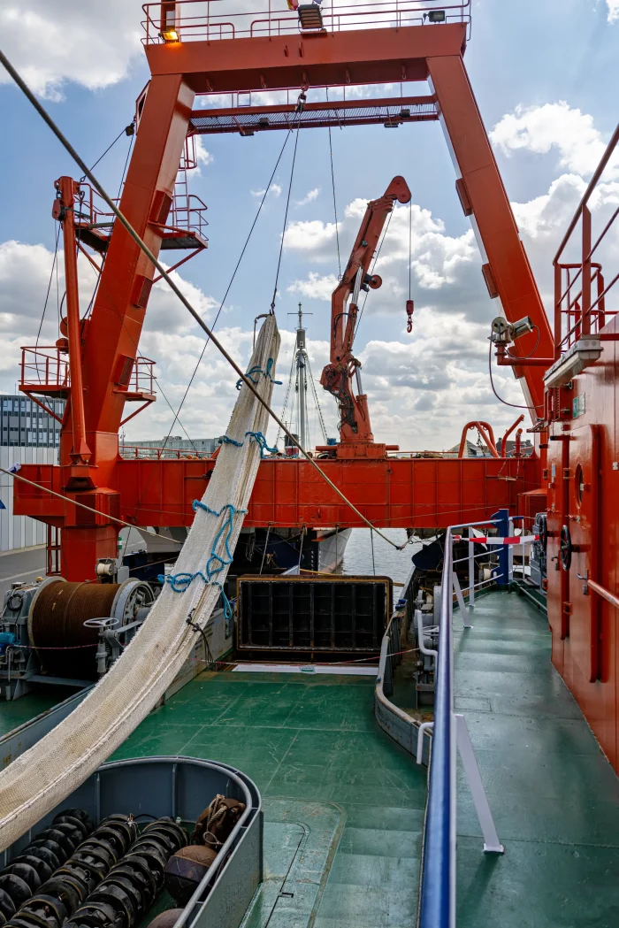 Deck eines Forschungsschiffs mit großen orangenen Kränen und aufgewickelten Netzen unter bewölktem Himmel