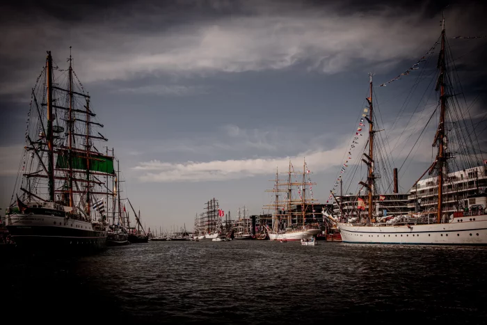 Mehrere große Segelschiffe mit hohen Masten und gesetzten Segeln liegen im Hafen unter bewölktem Himmel