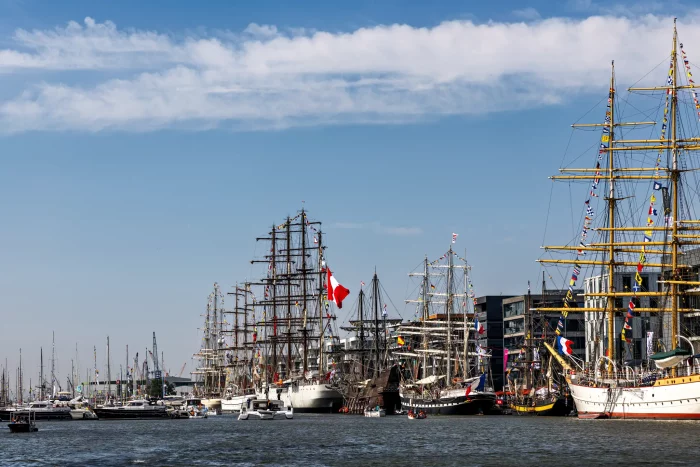 Mehrere große Segelschiffe mit hohen Masten und Flaggen liegen im Hafen unter blauem Himmel