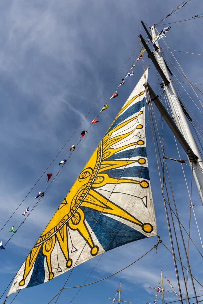 Segel mit gelbem und blauem Sonnenmotiv an einem Segelschiff, Mast mit Signalflaggen vor blauem Himmel