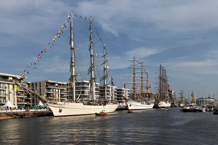 Drei große Segelschiffe mit hohen Masten und Flaggen liegen an einem Hafen mit Gebäuden im Hintergrund