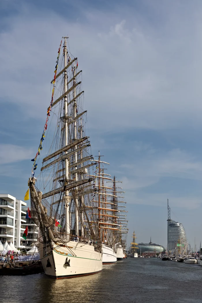 Segelschiffe mit hohen Masten und Flaggen am Hafen vor moderner Stadtkulisse unter blauem Himmel