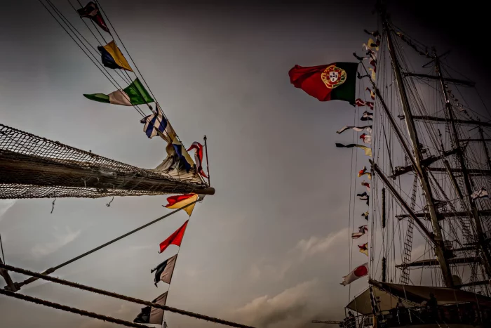Segelschiff mit Mast und zahlreichen Flaggen, darunter die Flagge Portugals, vor bewölktem Himmel