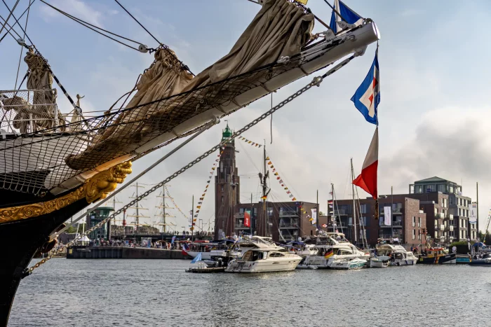 Vorderer Teil eines Segelschiffs mit Flaggen, im Hintergrund Hafen mit Yachten und einem hohen Leuchtturm