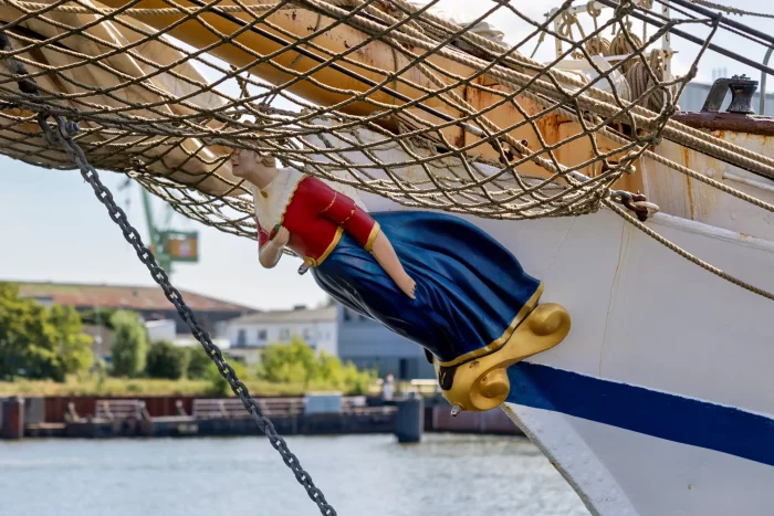 Schiffsfigur einer Frau in rotem Oberteil und blauem Rock am Bug eines Segelschiffs