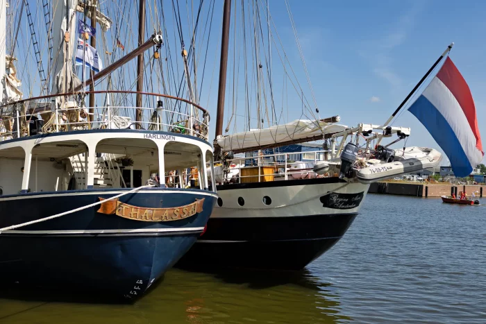 Zwei Segelschiffe nebeneinander im Hafen, eines mit niederländischer Flagge am Heck