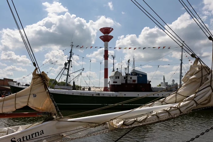 Segelschiff 'Saturna' mit getakeltem Mast im Hafen, dahinter ein Frachtschiff und ein hoher rot-weißer Sendeturm unter bewölktem Himmel