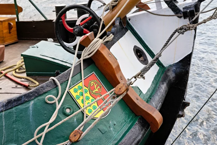 Bug eines Segelschiffs mit grüner Bordwand, roten und schwarzen Steuerrad und einem Wappen mit gelbem Baum und roten Elementen
