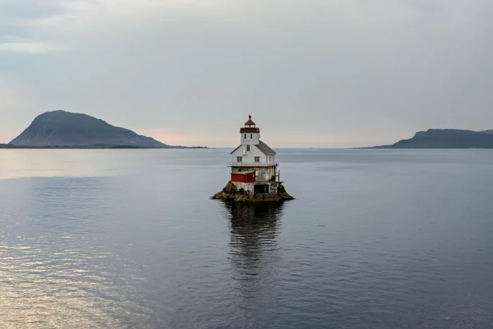Leuchtturm auf kleinem Felsen im ruhigen Meer, im Hintergrund zwei bewaldete Inseln unter bewölktem Himmel
