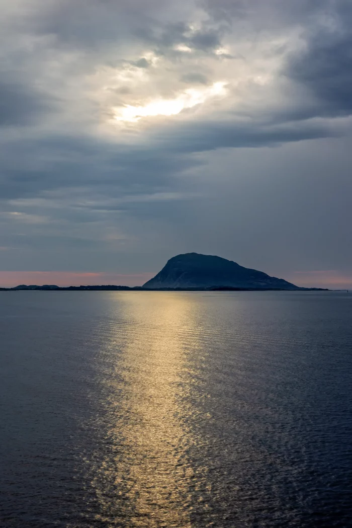 Sonnenlicht spiegelt sich auf ruhiger Wasseroberfläche vor einer Insel mit Hügel unter bewölktem Himmel