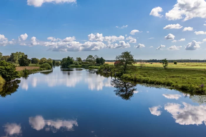 Flusslandschaft mit ruhigem Wasser, das Wolken und Bäume spiegelt, unter blauem Himmel im August