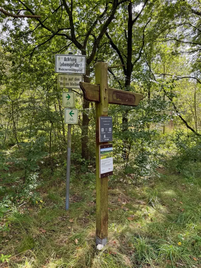 Wegweiser aus Holz mit der Aufschrift 'Buhlweg', umgeben von Bäumen und Gras. Links ein Warnschild 'Achtung Lebensgefahr!' und darunter zwei grüne Symbole für Wander- und Radwege.