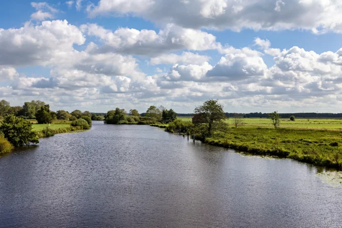 Eine ruhige Landschaft mit einem gewundenen Fluss, umgeben von grasbewachsenen Ufern und Bäumen. Der Himmel ist teilweise bewölkt, mit einem Hauch von Blau, der durchschaut. Ein kleines rotes Gebäude ist in der Ferne nahe dem Wasserrand zu sehen.