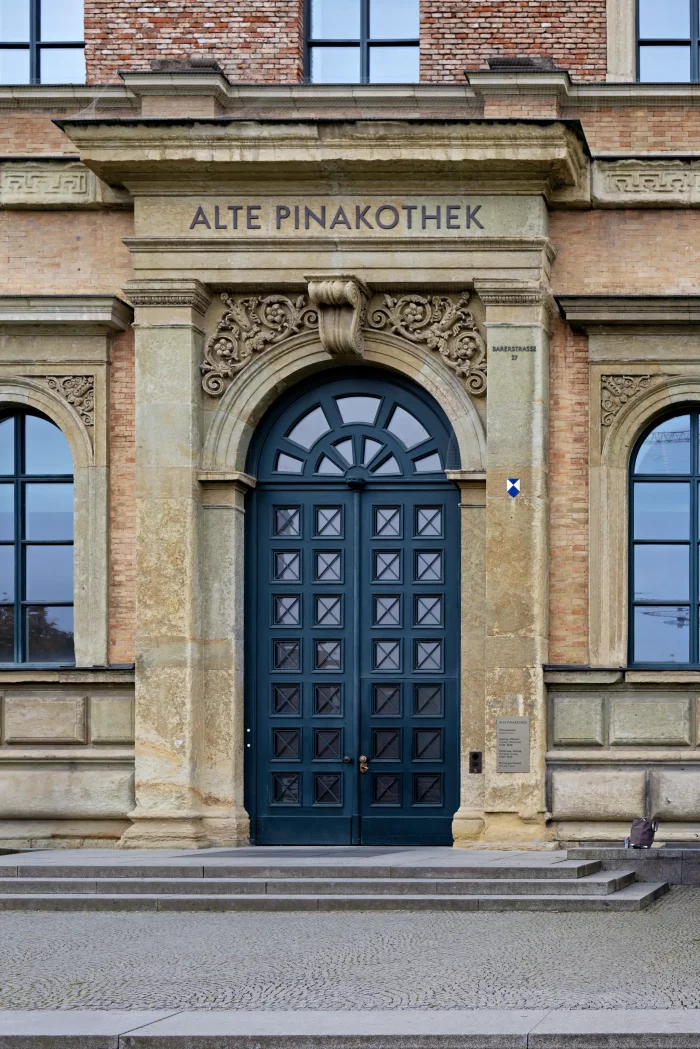 Fassade der Alten Pinakothek mit verziertem Sandsteintor und dunkelblauer Holztür mit Fenstern.