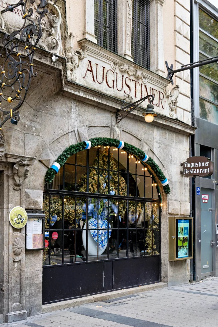 Fassade des Augustiner Restaurants in München mit großem Bogenfenster, geschmückt mit grün-weiß-blauem Girlandenkranz und Wappen im Fenster.