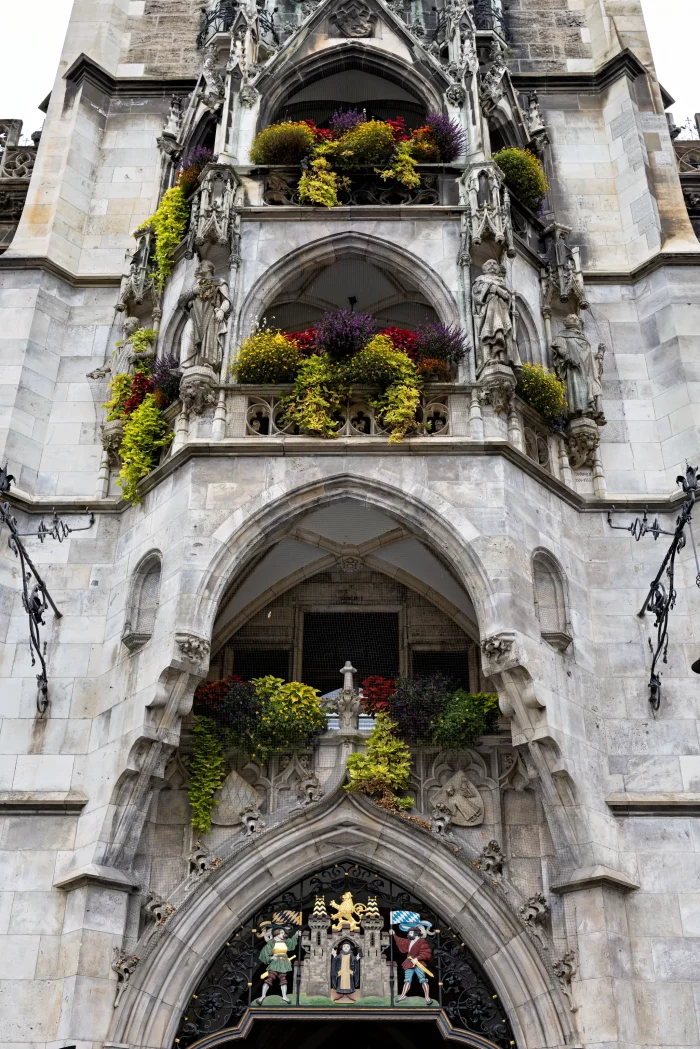 Fassadendetail des Münchner Rathauses am Marienplatz mit gotischen Bögen, Steinfiguren und bunten Blumenarrangements in mehreren Ebenen.