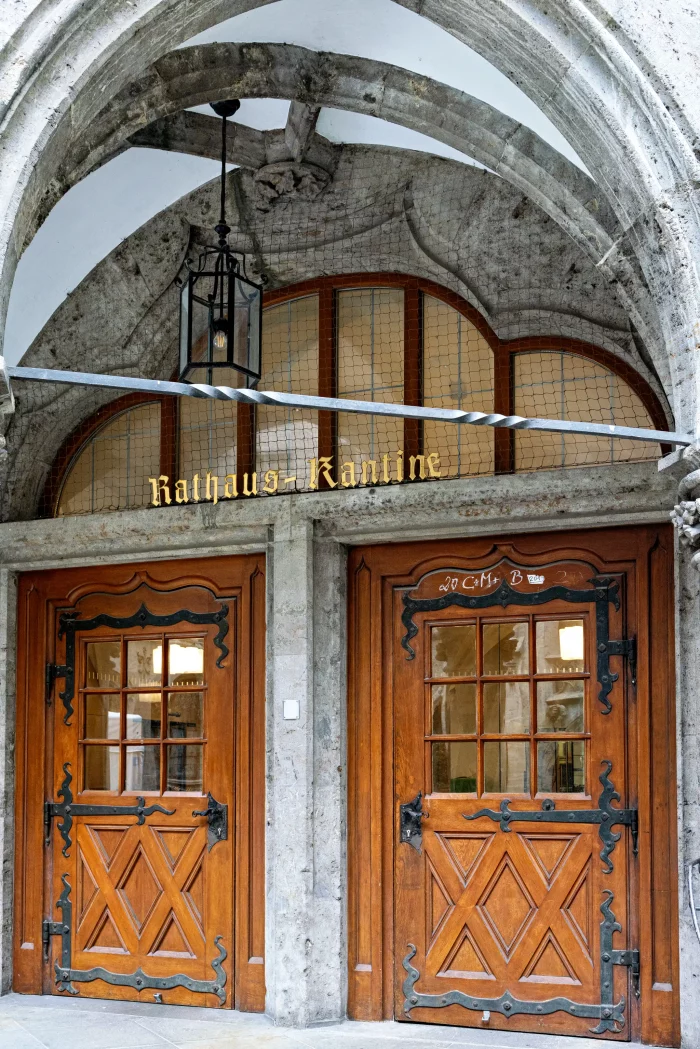 Holztüren mit Fenstereinsätzen und schmiedeeisernen Beschlägen unter einem steinernen Bogen mit dem Schriftzug 'Rathaus-Kantine' in goldenen Buchstaben