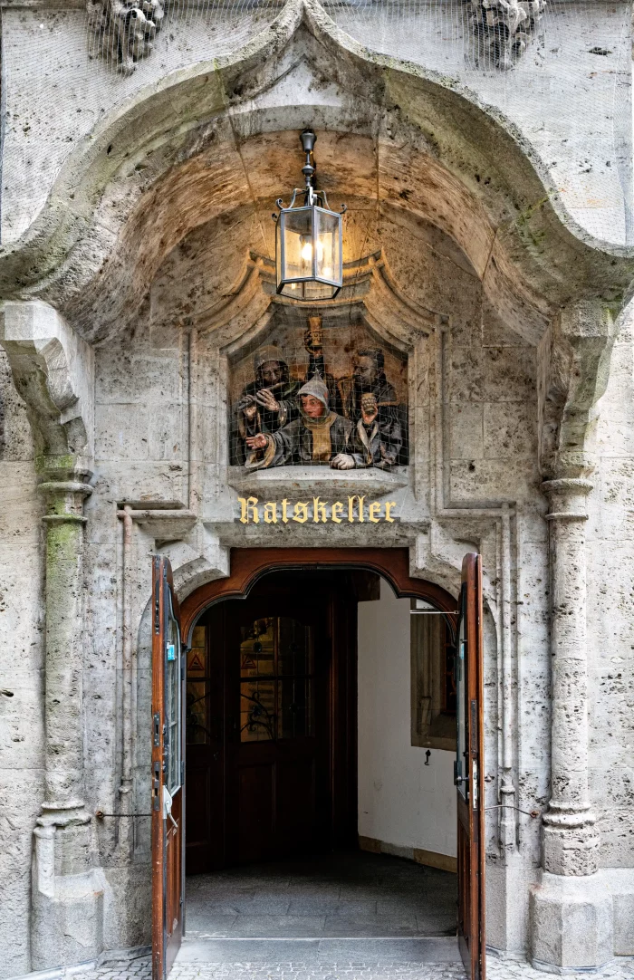 Offener Eingang zum Ratskeller am Münchner Marienplatz mit steinerner, verzierten Portalumrahmung und einer Laterne darüber.