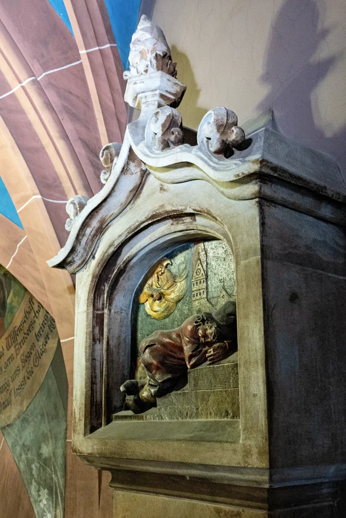 Steinrelief an der Wand im Münchner Rathaus, zeigt eine liegende Figur auf Treppen mit einem Engel und einem Turm im Hintergrund.