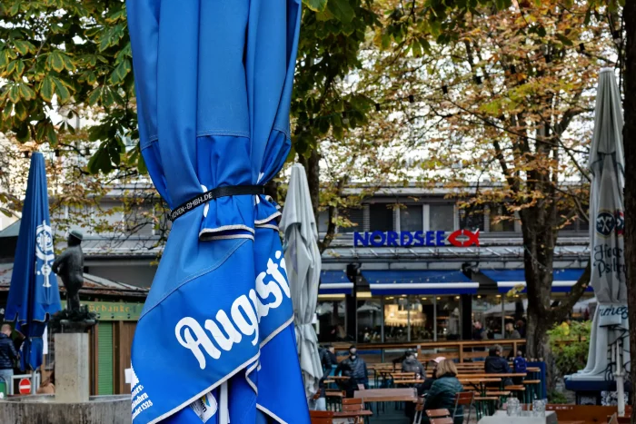 Geschlossene blaue Augustiner-Sonnenschirme vor dem Nordsee-Restaurant am Viktualienmarkt in München, umgeben von herbstlichen Bäumen.
