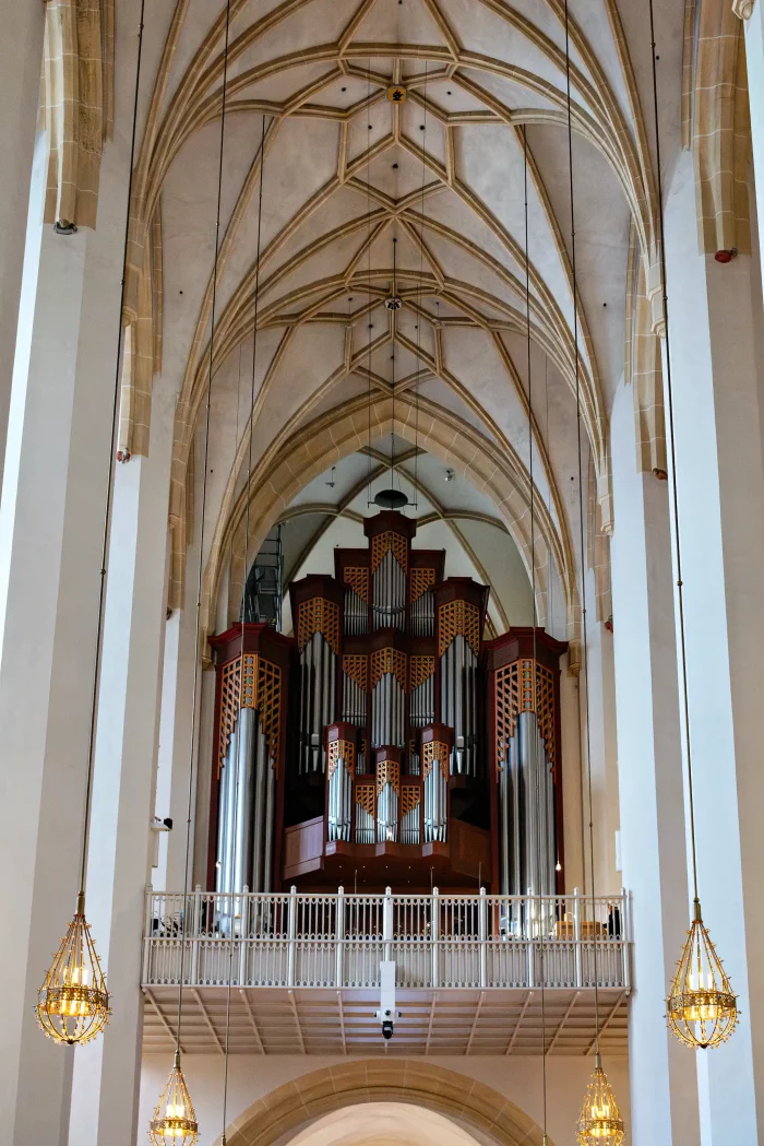Gotische Gewölbedecke und große Orgel mit silbernen Pfeifen in der Frauenkirche München, Blick auf die Empore mit weißen Geländern und goldenen Kronleuchtern.